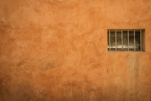 An old, barred rectangle window on the right side of an orange weathered plaster wall.