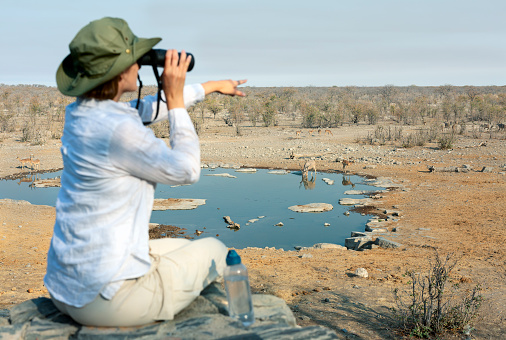 Rear view of a young blond woman on safari sitting on the rock looking through binoculars - travel concept