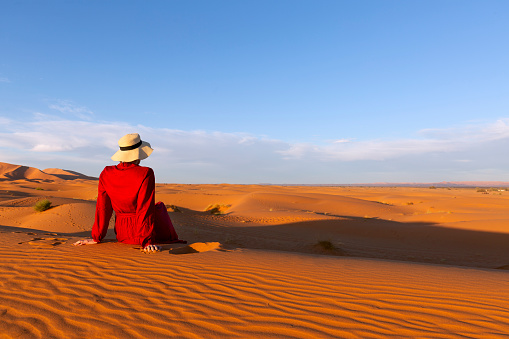 A young woman dressed in red and hat sitting on the desert dunes