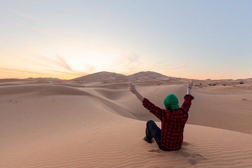 A young man  with open arm sitting on the desert dunes - travel concept