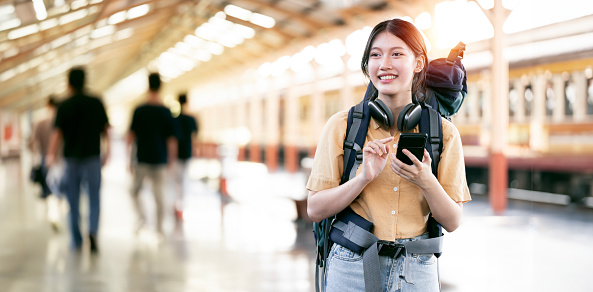 Enjoying travel. Young pretty woman with backpack standing on railway platform at train station and using smartphone.