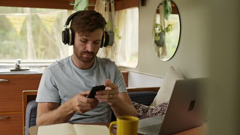 Man, headphones and freelancer streaming song on mobile app, playlist and typing on smartphone for communication. Male person, laptop and remote work to jazz, internet and relax on caravan adventure