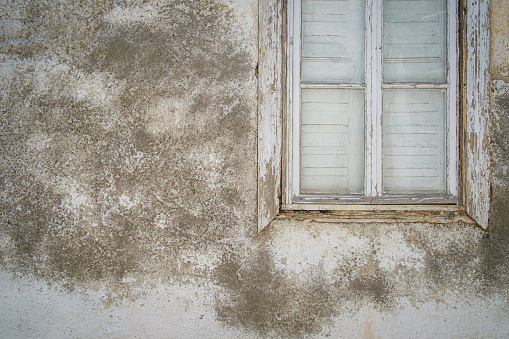 A peeling old window with white shutters on a dirty stucco wall.