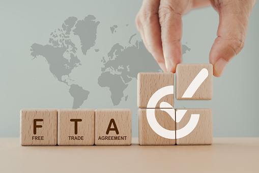 Hand arranged wooden cube blocks with approved signed and word FTA,  abbreviation for Free Trade Agreement, and blurred stack of coin on world map background