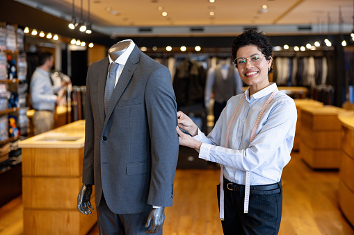Happy seamstress sewing a business suit on a mannequin and looking at the camera smiling - clothing store concepts