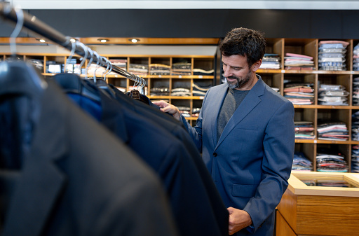Happy Latin American elegant man buying a suit at a clothing store and looking at jackets hanging on a rack