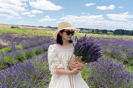 A woman clad in a white summer dress and wearing a straw hat and sunglasses stands in a lavender field, holding a bouquet of freshly picked lavender. The image captures a clear sky with scattered clouds above and a serene, natural landscape in the background.