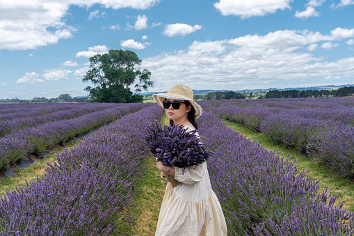 A woman clad in a white summer dress and wearing a straw hat stands in a lavender field, holding a bouquet of freshly picked lavender. The image captures a clear sky with scattered clouds above and a serene, natural landscape in the background.