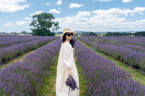A woman clad in a white summer dress and wearing a straw hat stands in a lavender field, holding a bouquet of freshly picked lavender. The image captures a clear sky with scattered clouds above and a serene, natural landscape in the background.