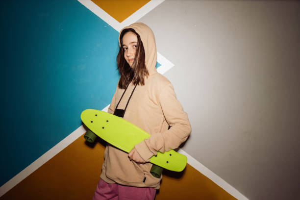 Girl in hooded sweatshirt holding skateboard and looking at camera against colored wall at home