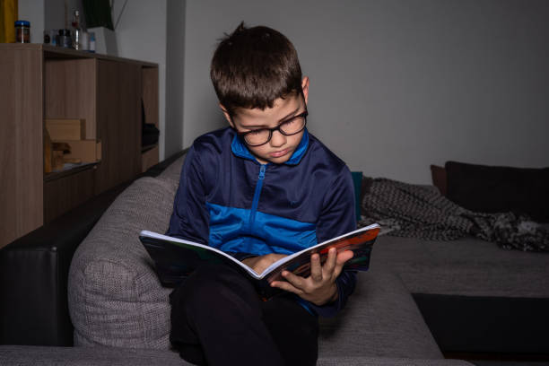 Cute boy with reading glasses sitting on sofa and reading a book at home