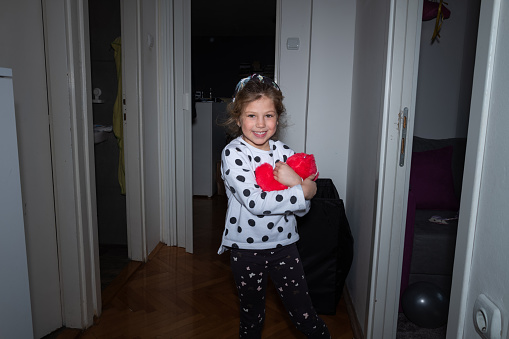 Cute little girl standing and embracing red soft toy in the shape of heart at home, smiling and looking at camera
