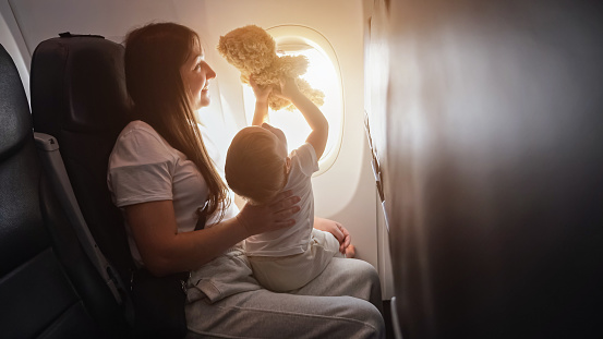 Mom takes flight and cradles toddler girl in arms. Toddler daughter joyfully plays and raises up toy bear during journey, sunlight