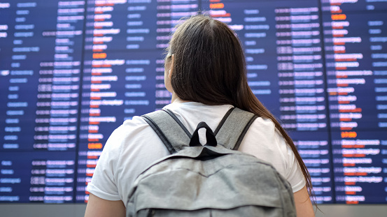 Woman stands at airport and looks at flight schedule hanging on wall. Lady searches for information about flight eager to find departure gate