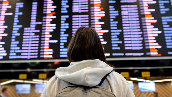 Female stands at airport terminal timetable looking for flight schedule on wall. Brunette lady searches for data about flight eager to get departure gate