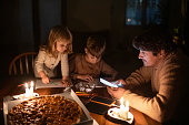 Family sitting at home with candles and eating pizza during blackout. Children are drawing, man is using solar power bank to charge his smartphone. Energy crisis concept.