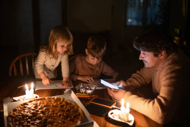 Family sitting at home with candles and eating pizza during blackout. Children are drawing, man is using solar power bank to charge his smartphone. Energy crisis concept. stock photo