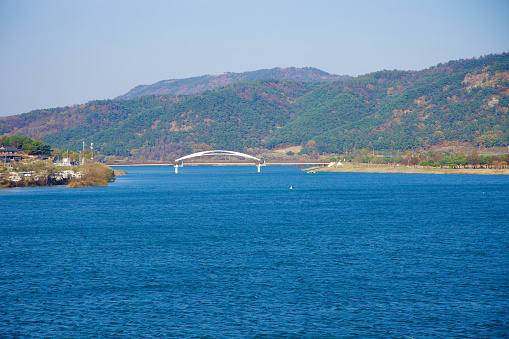 Sangju City, South Korea - November 18th, 2023: Looking up the Nakdong River from Sangju Weir, the distinctive double-leaping metal awnings of Gyeongcheon Bridge lead to Gyeongcheon Island Park, with mountains framing the background.