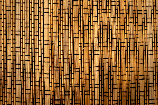 Surface of a naturally woven brown bamboo mat interwoven with dark threads, close-up.  Background texture