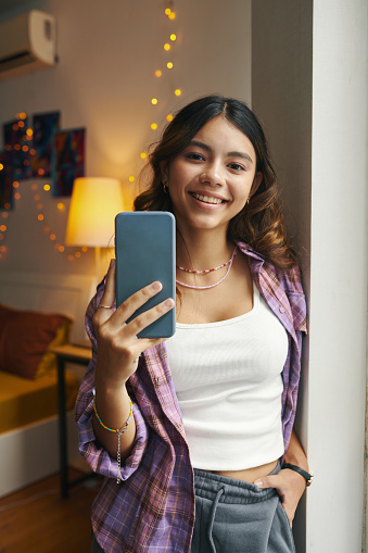 Portrait of cheerful teenage girl in loungewear standing at bedroom window when taking selfie