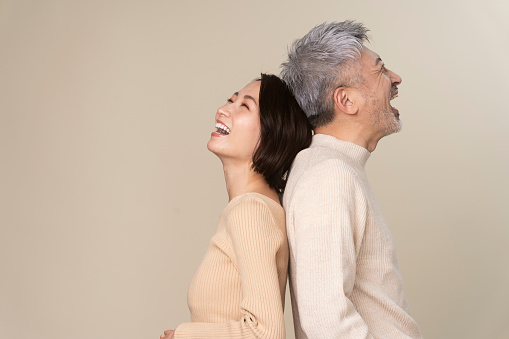 A middle-aged Japanese couple stands back to back.They are laughing.Studio shot  on beige background.