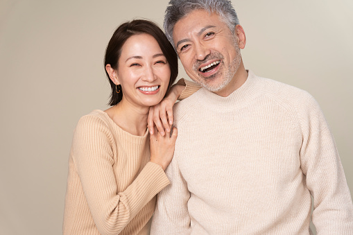 A middle-aged Japanese couple is smiling at the camera.They stands side by side. A wife puts her hand on her  husband's shoulders.