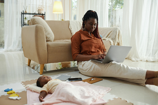 Black woman sitting on floor and working on laptop when her newborn daughter sleeping near by