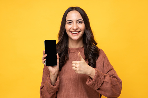 Photo of beautiful happy smiling young woman wearing sweatshirt standing isolated over yellow background showing mobile phone with blank screen for camera