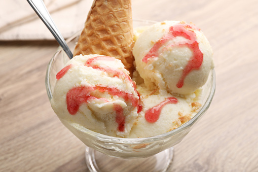 Delicious scoops of vanilla ice cream with wafer cone and strawberry topping in glass dessert bowl on wooden table, closeup