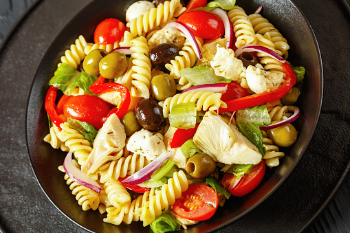 fusilli salad with mini mozzarella, artichoke, tomatoes, romaine lettuce, olives, onion, bell pepper and seasoning in black bowl on black wooden table, close-up