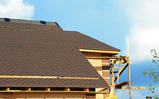 A close-up of an unfinished roofing construction with asphalt roofing shingles installation on the ridge of the rooftop and roofing facia boards and soffit uninstalled.