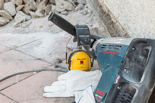 Protective ear muffs. Equipment for personal protection (DPI – PPE) of the famous 3M Company, brand Peltor. American multinational operating in more than 70 countries in the fields of industry, worker safety, healthcare and consumer goods. To the side, a Bosch jackhammer