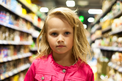 Portrait of cute child girl 5 year old at shelves with groceries in store background, serious looking at camera. Kid girl shopping buying in supermarket. Retail shopping concept. Copy ad text space