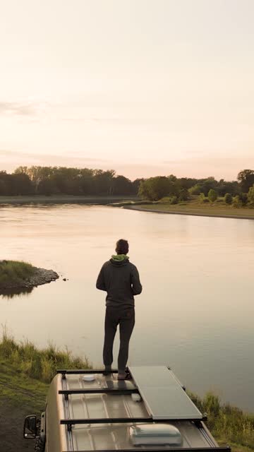 Man Standing on a Camper Van Next to a River at Sunset