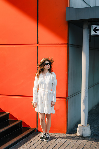 Woman in white dress and sunglasses on the background of orange wall lit by sunset light