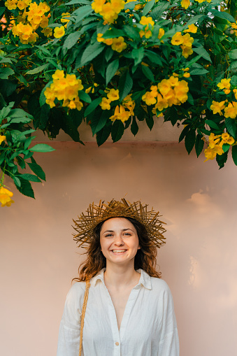 Portrait of woman in straw hat standing under the bush with yellow flowers on the background of the wall