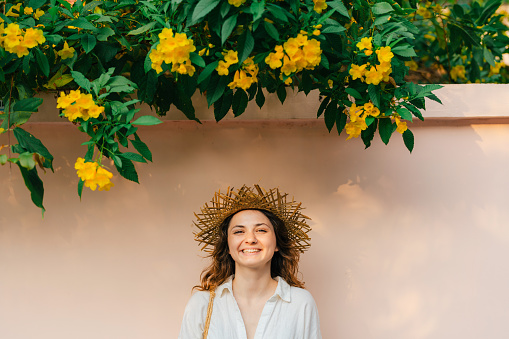 Portrait of woman in straw hat standing under the bush with yellow flowers on the background of the wall