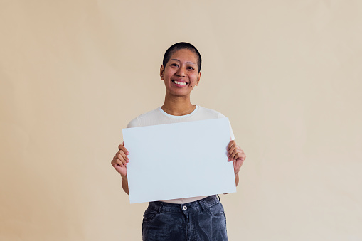 Young woman standing in front of a cream coloured background. She is looking at the camera while holding a blank sign.