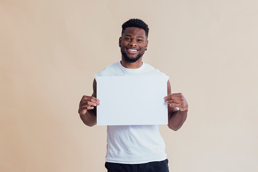 Young man standing in front of a cream coloured background. He is looking at the camera while holding a blank sign.