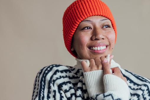Young woman standing in front of a cream coloured background. She is looking away from the camera wearing an orange beanie and striped jumper.