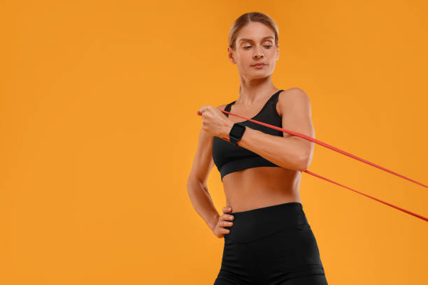 istock Woman exercising with elastic resistance band on orange background, low angle view. Space for text 1954485011
