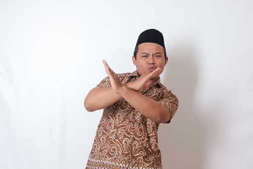 Portrait of excited Asian man wearing batik shirt and songkok  showing X sign of hands, refusing an invitation from someone, making prohibitive gesture. Isolated image on gray background