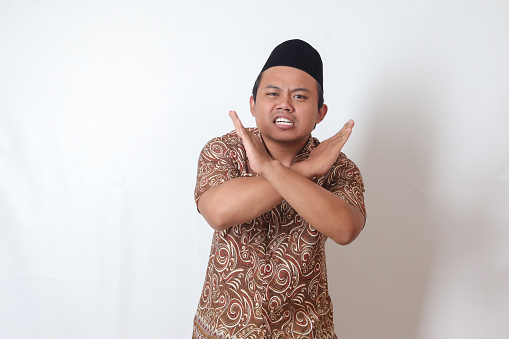 Portrait of excited Asian man wearing batik shirt and songkok  showing X sign of hands, refusing an invitation from someone, making prohibitive gesture. Isolated image on gray background