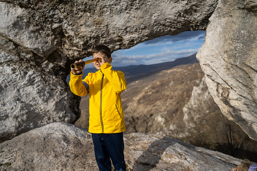Boy looking through telescope while standing by stone arch on rocky mountain