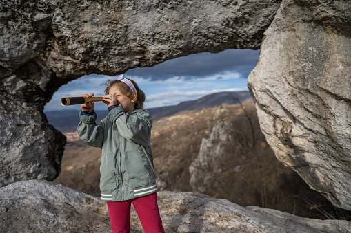 Little girl looking through telescope while standing by stone arch on rocky mountain