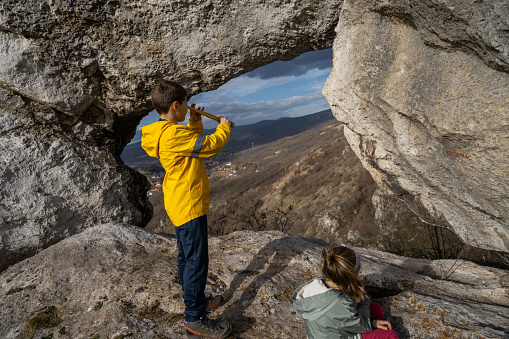 Boy looking through telescope by stone arch while sister sitting next to him on rocky mountain