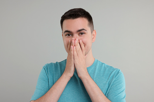 Embarrassed man covering mouth with hands on grey background