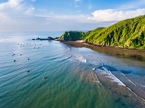 A serene beach bordered by lush green hills. The ocean is calm with gentle waves lapping at the shore. Several boats are scattered in the water near the shore. The sky is clear and blue, indicating fair weather. The perspective from above provides a comprehensive view of the landscape. Shot taken on Lombok.