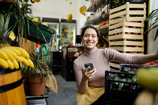 Portrait of a young woman working at the grocery store, using smart phone and arranging fruits and vegetables.