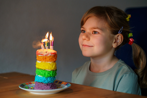 Happy little preschool girl celebrating birthday. Closeup of child with homemade rainbow cake, indoor. Happy healthy toddler blowing six candles on cake. Selective focus on cake.